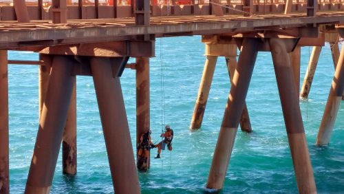 Two rope access technicians are suspended from a jetty, performing maintenance on its underside. The large cylindrical pillars of the jetty rise from the turquoise sea, supporting a wide platform above. The sunlight illuminates the water and part of the jetty, highlighting the technicians equipped with orange safety gear, clearly focused on their tasks.
