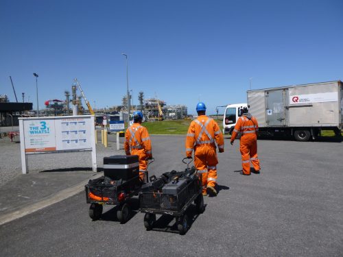 Three Vertech Group employees, dressed in bright orange high-visibility jackets and blue helmets, walk towards an industrial facility with equipment in tow. They are passing a safety sign labelled 'The 3 What's?' outlining life-saving rules at the entrance to the plant. In the background, the expansive industrial complex features various metallic structures and machinery under a clear blue sky.