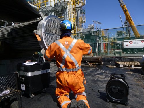 A Vertech Group technician, clad in an orange high-visibility jacket and blue helmet, operates a specialised inspection device at an industrial site. The technician is focused on examining a large piece of machinery using an Olympus inspection device connected to a laptop displaying the diagnostic data. The background features a complex array of yellow and grey industrial structures under a clear sky.