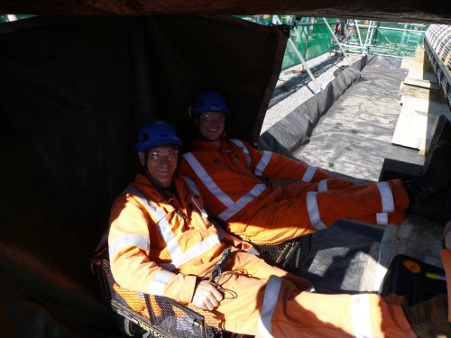 Two Vertech Group employees, wearing safety gear and helmets, sit comfortably inside a shaded, makeshift rest area at a construction site. They are dressed in bright orange high-visibility jackets and blue helmets, each marked with their names. The background reveals a partially visible construction setup under bright daylight.